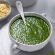bowl of green coriander chutney with a white spoon. There is sev, bhel and coriander leaves in the frame around the bowl.