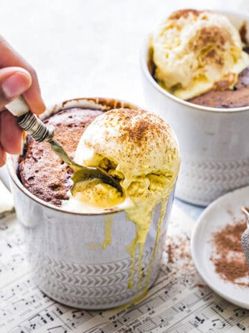 The image shows two mugs of Nutella mug cake topped with a generous scoop of vanilla ice cream. A lady is digging into the cake with a spoon, ready to enjoy the delicious treat. The white mug adds a clean, cozy feel to the scene, highlighting the indulgent dessert.