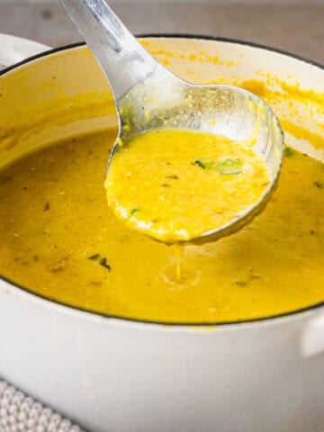 Ladleful of red lentil dal soup and a white pot of soup.