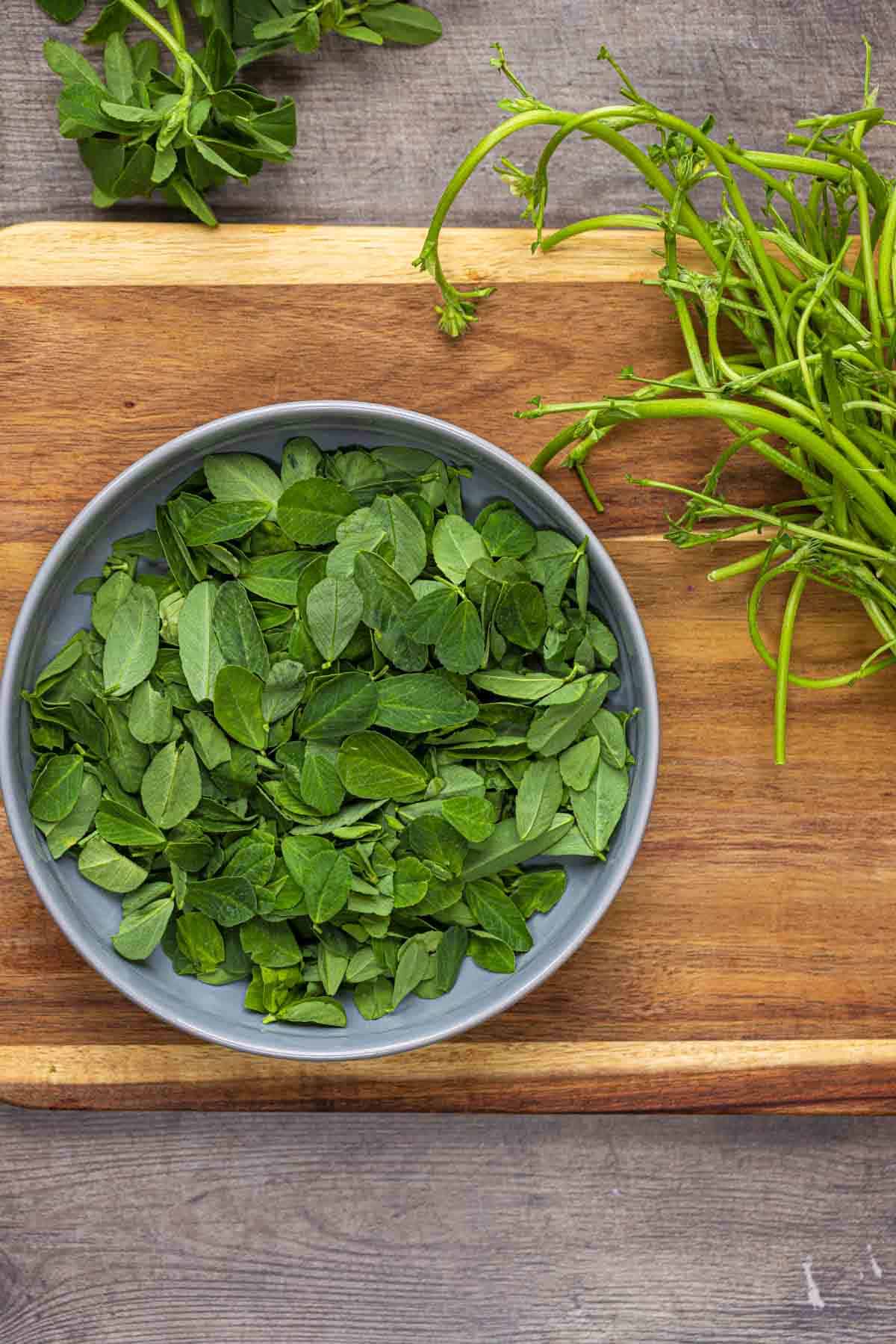 picked fresh methi leaves from the stem. the are in a grey bowl placed on a wooden board.