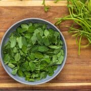 picked fresh methi leaves from the stem. the are in a grey bowl placed on a wooden board.