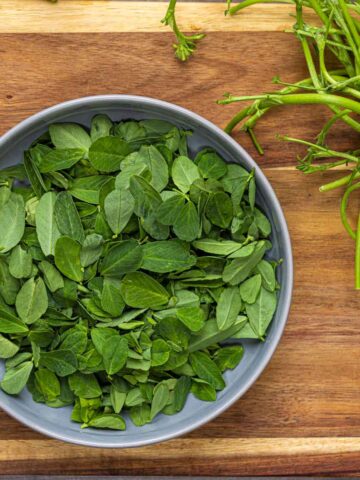 picked fresh methi leaves from the stem. the are in a grey bowl placed on a wooden board.