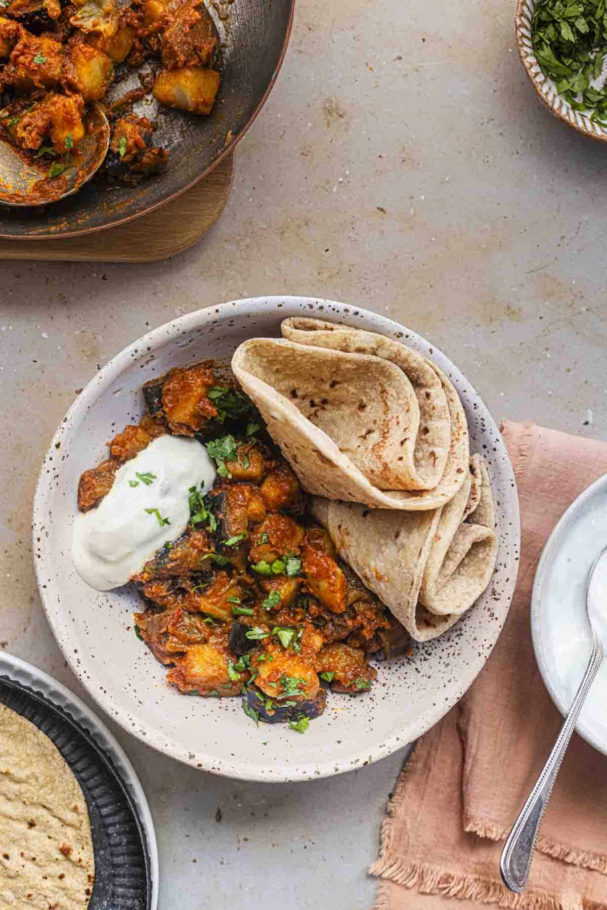 aloo baingan served with chapatis and cucumber raita in a white bowl.