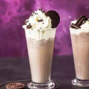 two glasses of cookies and cream milkshake topped with whipped cream and oreo cookies.