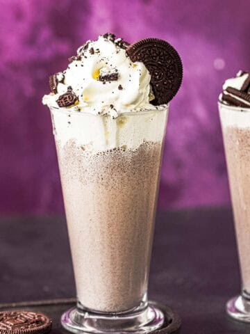two glasses of cookies and cream milkshake topped with whipped cream and oreo cookies.
