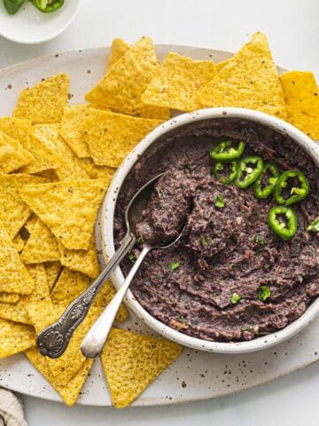 a white bowl of vegan black bean dip topped with slices of jalapeños and served with tortilla chips.