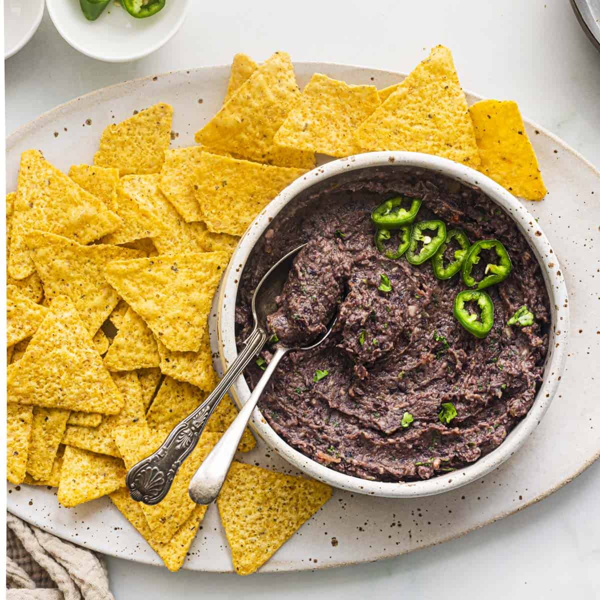 a white bowl of vegan black bean dip topped with slices of jalapeños and served with tortilla chips.